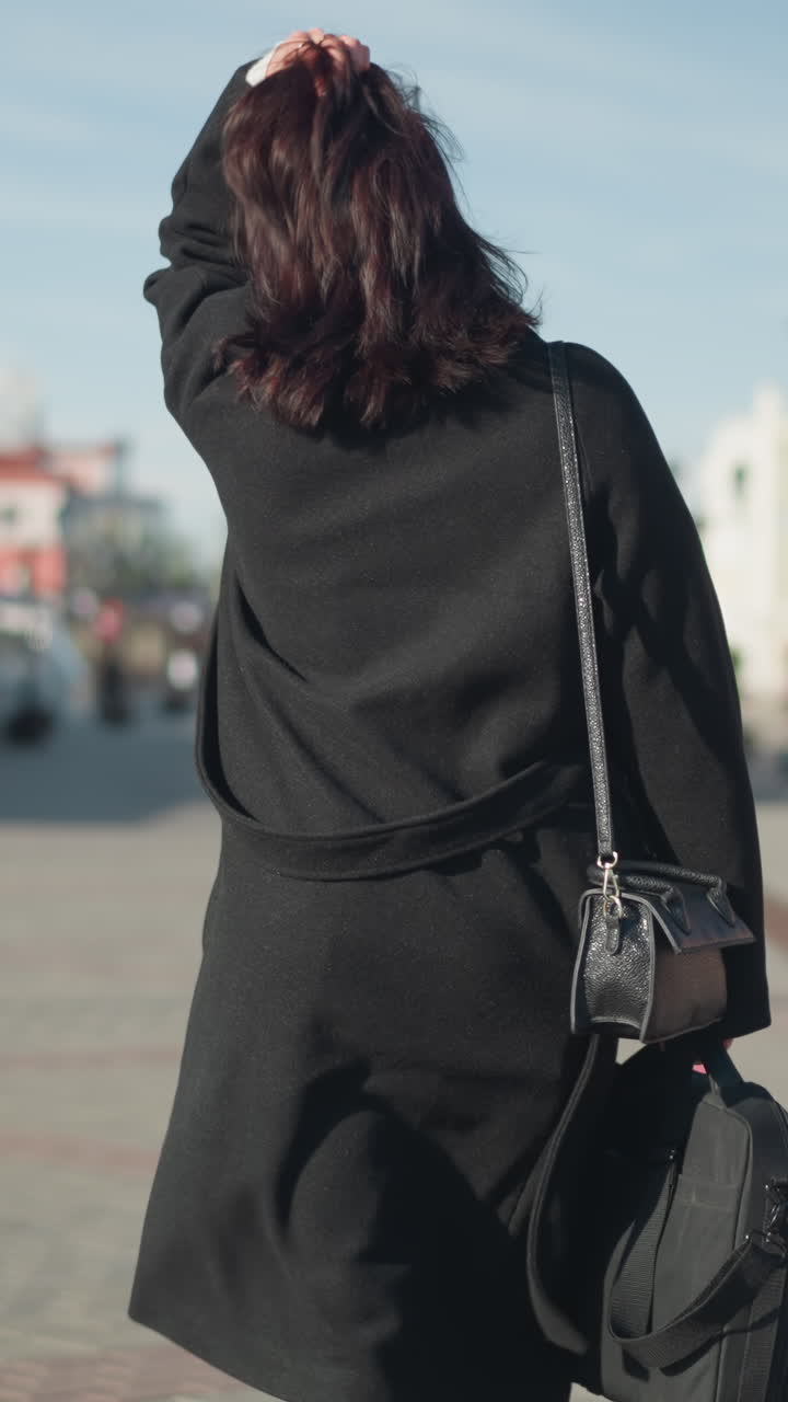 검은 코트를 입은 갈색 머리카락의 여인이 머리를 조정하고, 왼손에 세 개의 반지를 선보이며, 배경으로 주거 건물과 함께 은 도시 거리에 있습니다.