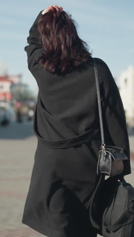 back view of a lady with brown hair in a black coat adjusting her head, showcasing three rings on her left hand on a sunny urban street with residential building with background