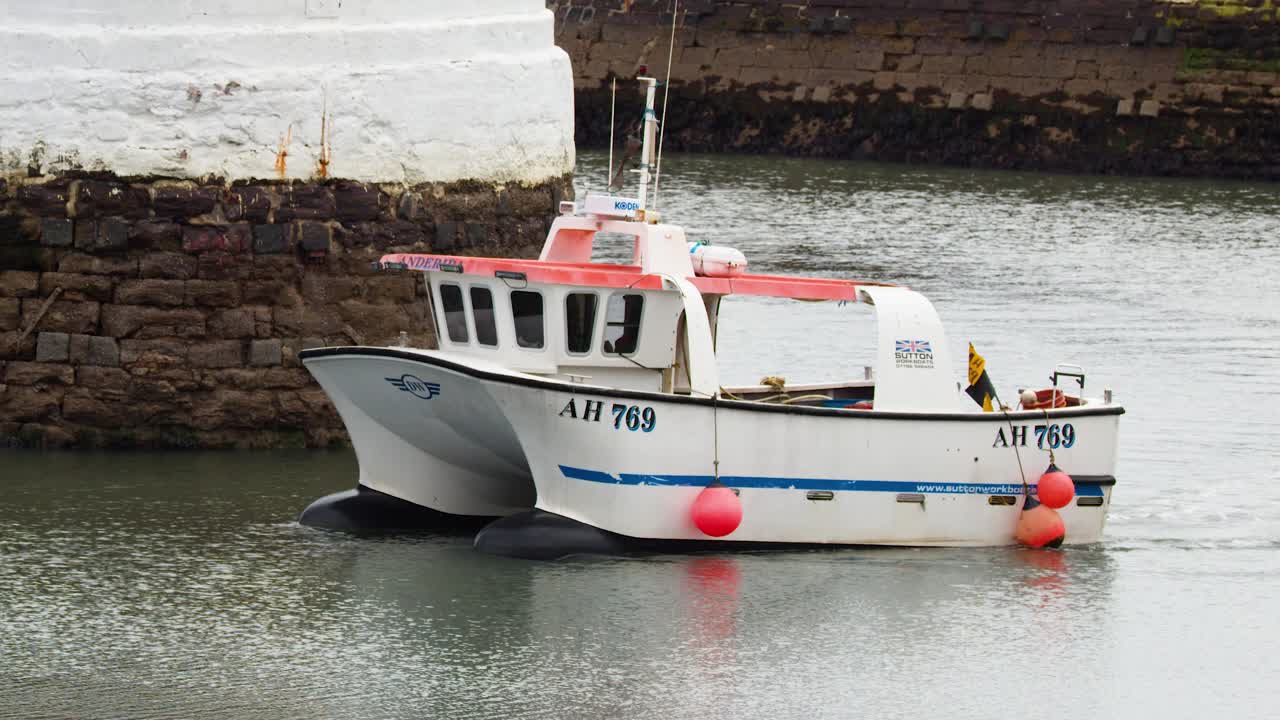 Small fishing boat slowly maneuvers past stone harbor wall in overcast daylight, steady camera angle