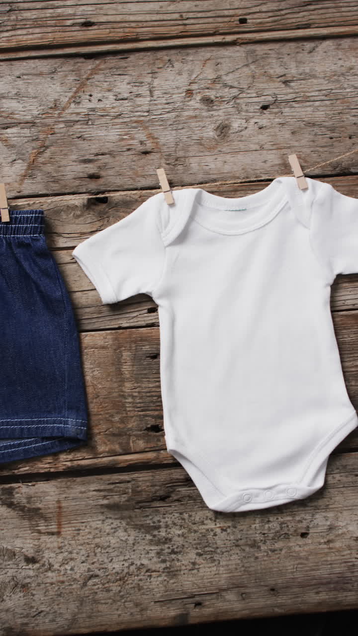 Vertical video of baby grow and denim shorts on clothes pegs with copy space on wooden background