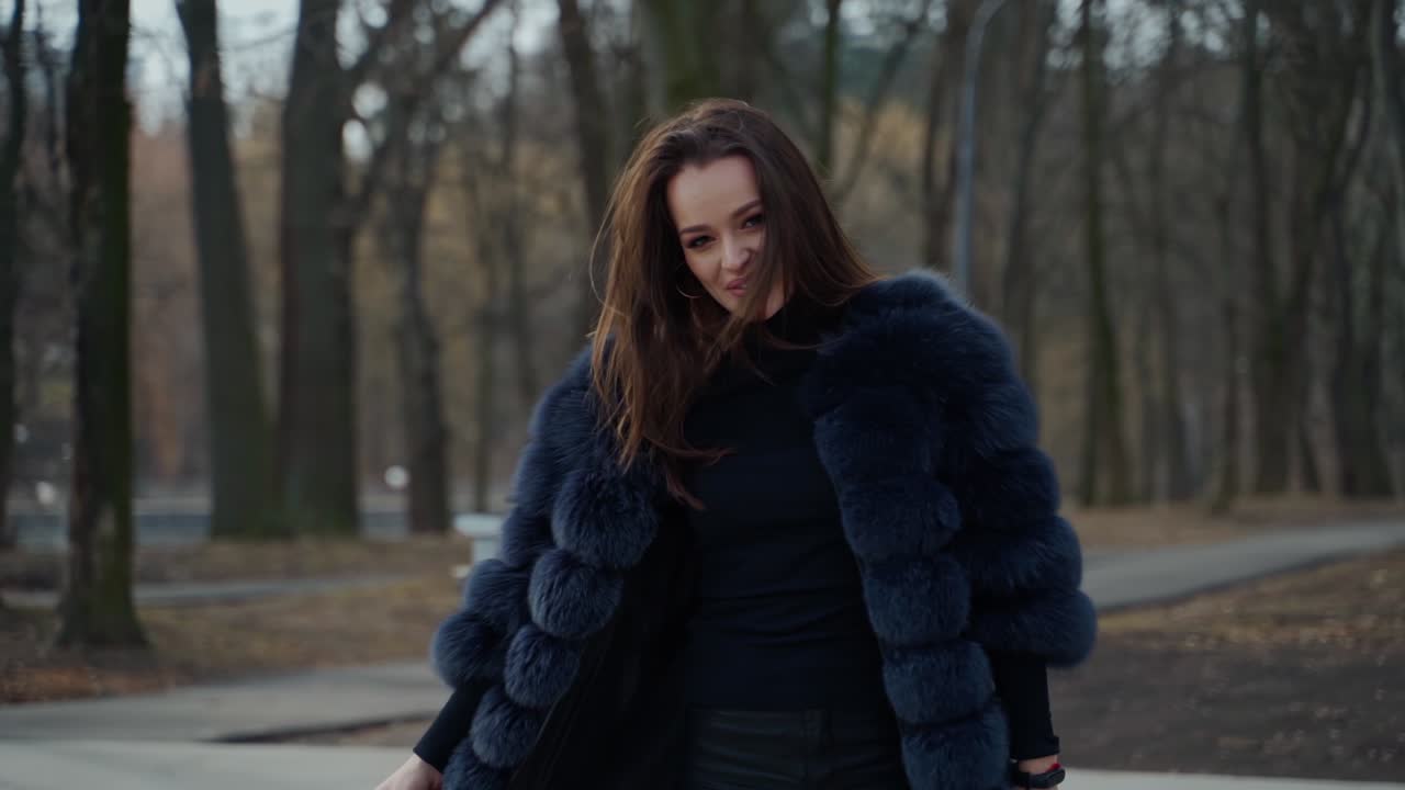 Glamour model posing on camera outdoors. Portrait of beautiful girl in a black fur coat smiling on the park background. Motion camera around.