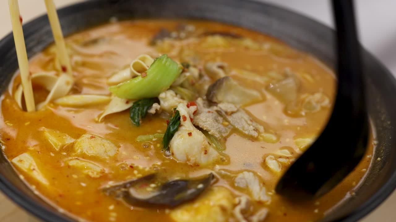 Hand uses chopsticks and spoon to lift pork, vegetables, and noodles from spicy hotpot soup
