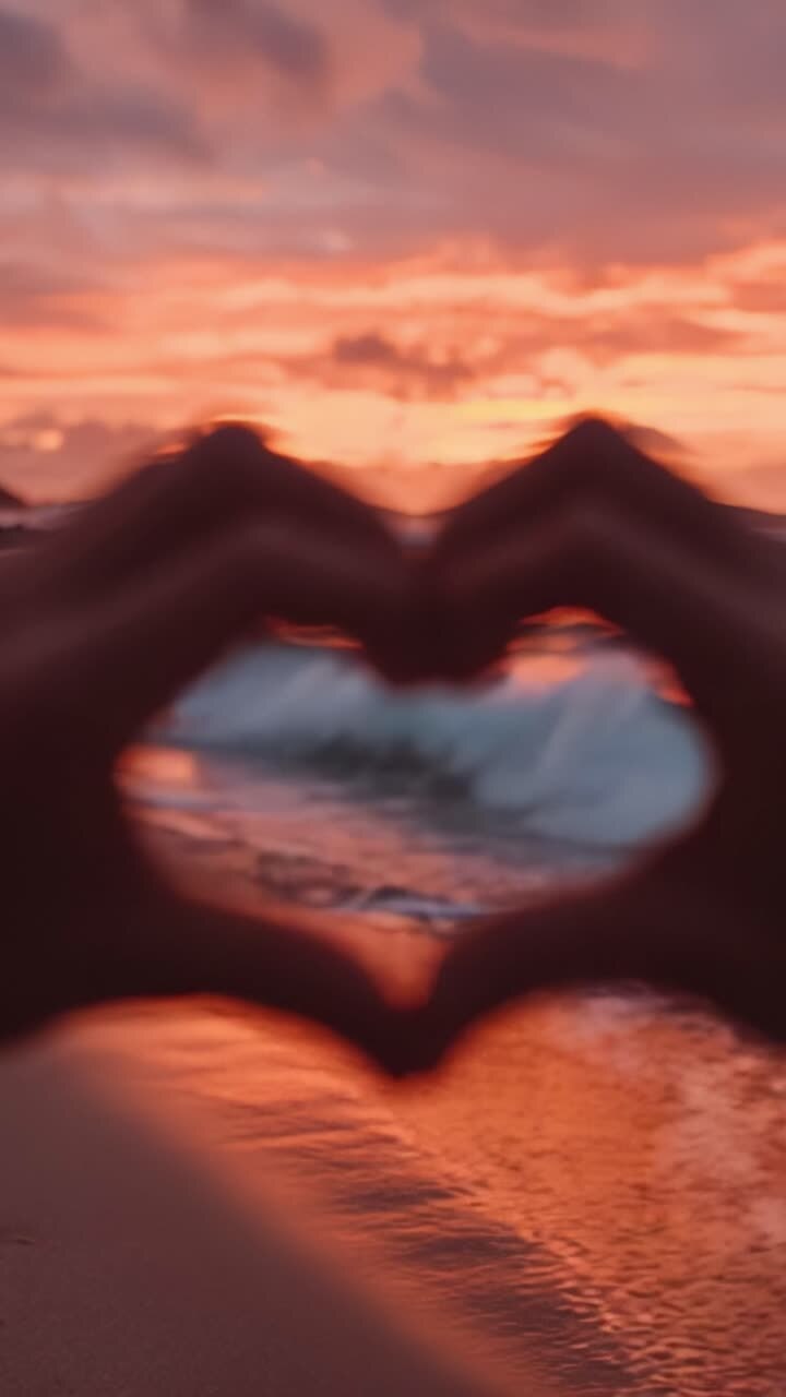Catturato in uno Splendido Tramonto: Mani che Formano un Cuore Contro lo Sfondo di Vivaci Onde Oceaniche e Colori Drammatici del Cielo al Crepuscolo