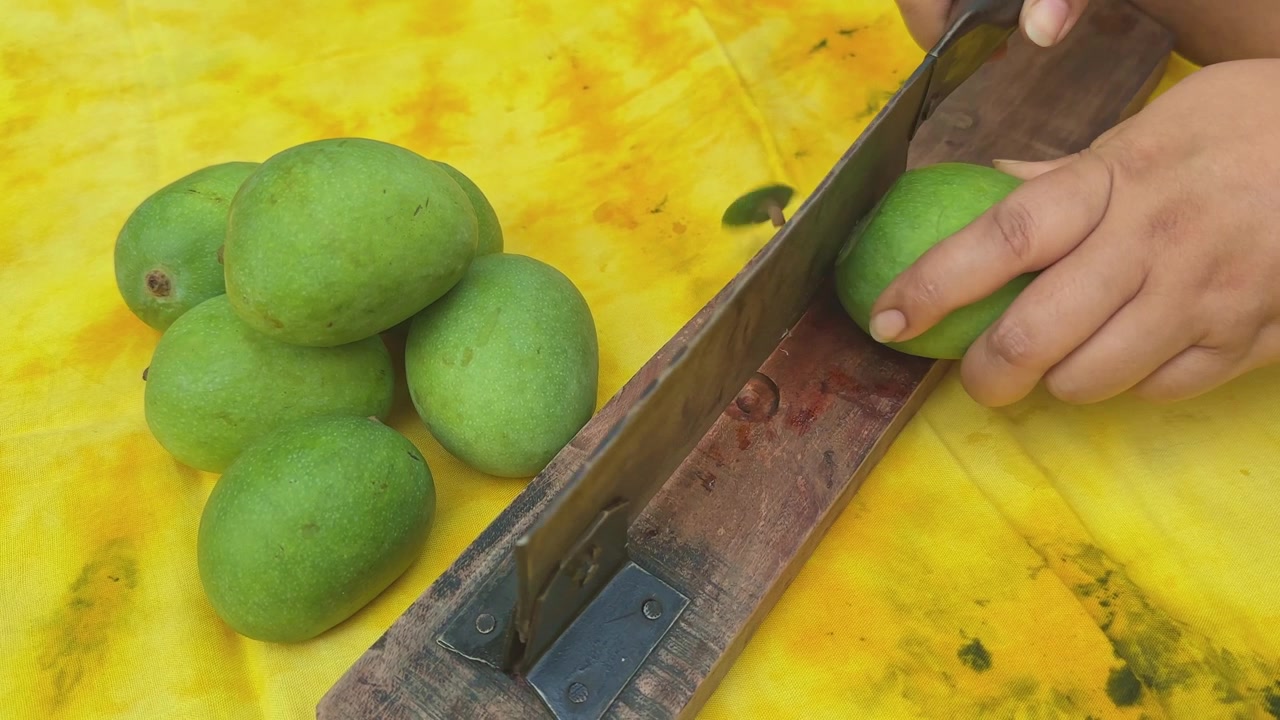 Vista ravvicinata di mani che usano un tradizionale taglia-mango indiano per rimuovere l'estremità del gambo e la linfa appiccicosa dai mango acerbi prima di affettarli per la preparazione di sottaceti fatti in casa