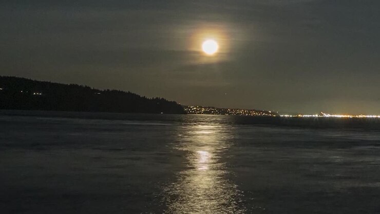 Hyperlapse con zoom indietro della luna piena che sorge sopra acqua scura e nera, luci della città e nuvole sfocate che scorrono.