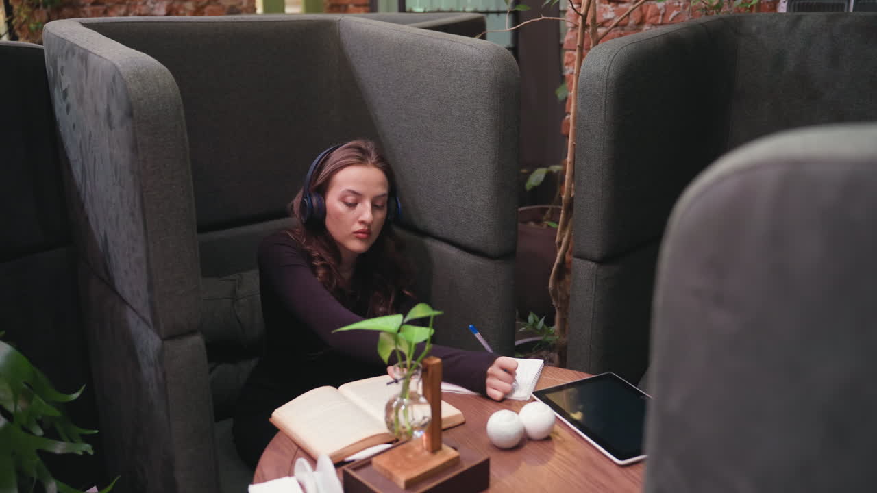 Top angle view of woman wearing headset sitting inside gray booth, writing in notebook while referencing open brown book on wooden table with tablet, plant, and two white spherical ornaments