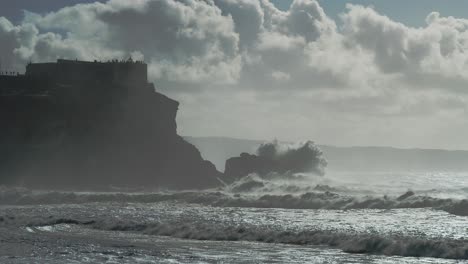 Wellen-Schlagen-Auf-Den-Felsen-Mit-Der-Festung-Farol-Da-Nazaré-Schöne-Filmische-Zeitlupenaufnahme