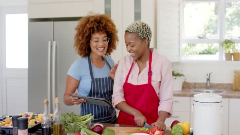 Happy-diverse-female-lesbian-couple-using-tablet-and-cooking-in-kitchen-in-slow-motion