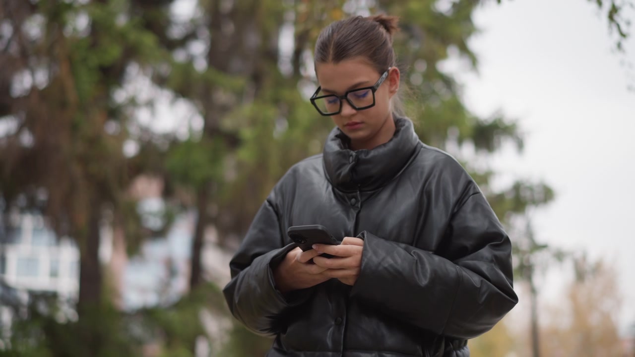 Primo piano di una giovane donna con occhiali e un piumino nero che tiene in mano il suo smartphone in un parco all'aperto con alberi e luce naturale soffusa. I suoi capelli sono raccolti in un piccolo chignon