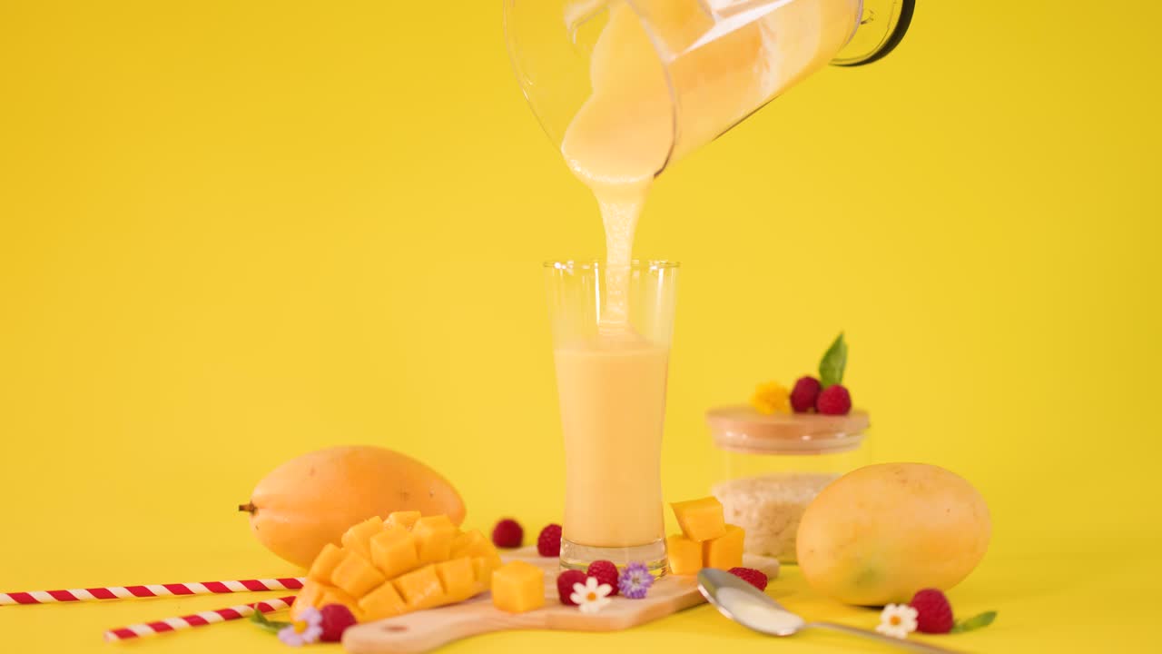 Thick mango smoothie pours into tall glass amid tropical fruit arrangement on bright yellow background
