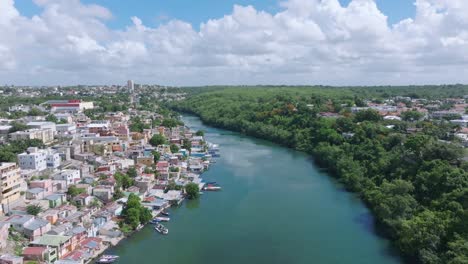 vuelo aéreo sobre el río rio romana con la ciudad de romana durante el día soleado - edificio en un lado y bosque en la otra costa