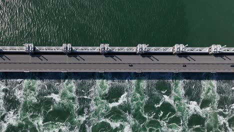 stunning wide aerial birdseye view of a storm surge barrier with a road and passing cars with calm water on one side and rough foamy water on the other