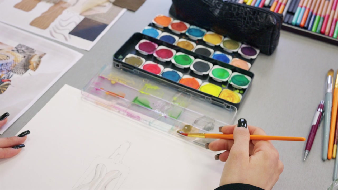 Overhead view of a fashion design workspace with a watercolor palette, colored pencils, brushes, black pencil case, fabric swatches, printed references