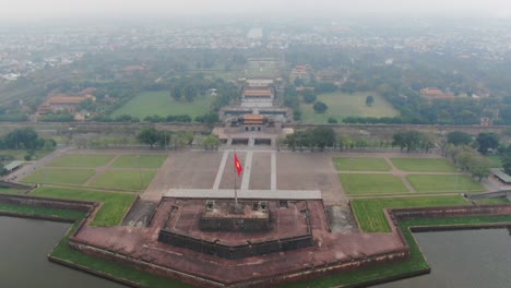 drone aerial view in vietnam flying over hue imperial stone brick fortress wall, green gardens, temples, buildings and flag on a cloudy and foggy day