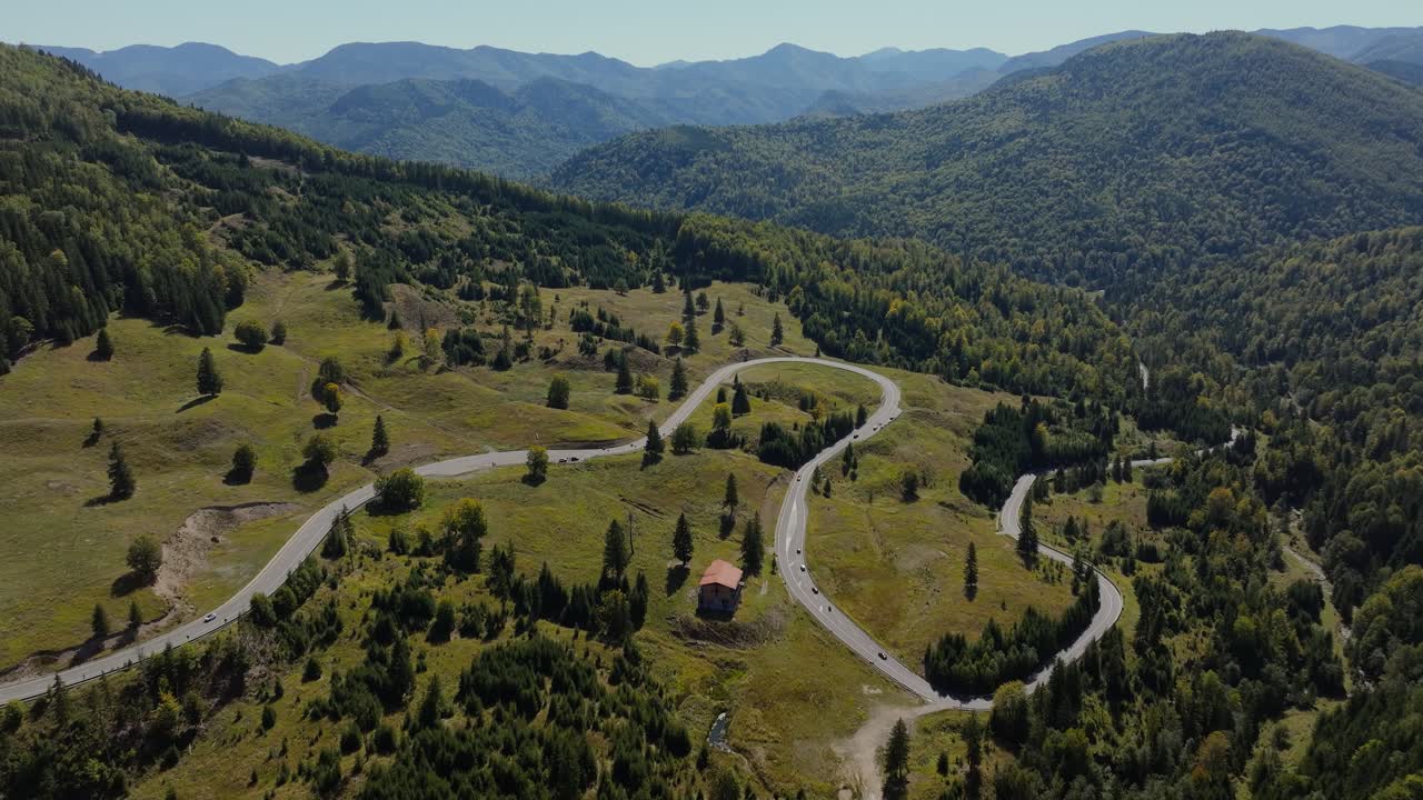 Wide aerial shot showing a winding mountain road weaving through expansive green hills and dense forest, distant mountain ranges under a clear blue sky