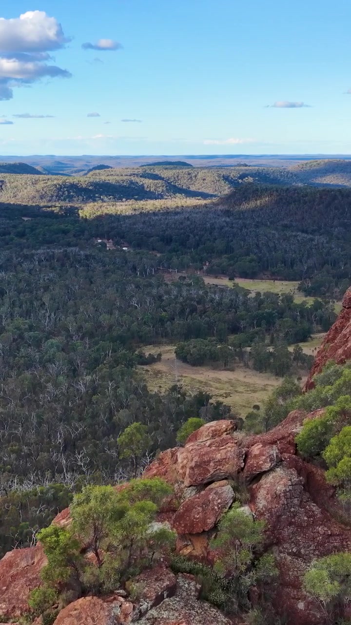 Un drone effettua una panoramica sull'affioramento rosso di Timor Rock, rivelando una vasta valle boscosa sotto una luce diurna soffusa.