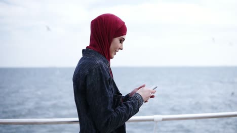 Una-Joven-Musulmana-Atractiva-Con-Hijab-En-La-Cabeza-Se-Desplaza-Y-Escribe-Algo-En-Su-Teléfono-Inteligente-Mientras-Camina-Cerca-De-La-Orilla-Del-Mar