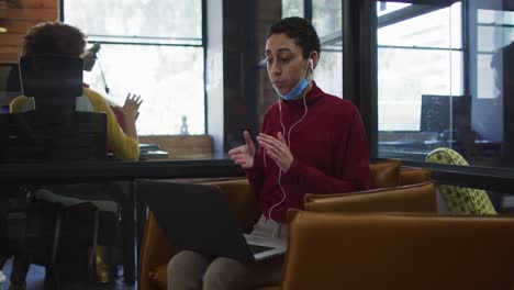 Caucasian-woman-with-lowered-face-mask-having-a-video-chat-on-laptop-at-modern-office