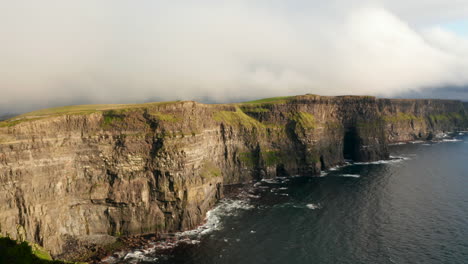 Aerial-ascending-footage-of-rocky-shore-lit-by-bright-sun.-High-vertical-rock-walls-above-rippled-sea-surface.-Cliffs-of-Moher,-Ireland