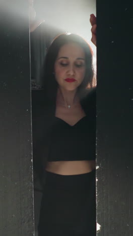 mujer elegante en traje negro y lápiz labial rojo parcialmente visible detrás de una pared de madera negra, cantando en luz suave mientras la cámara hace zoom dentro y fuera