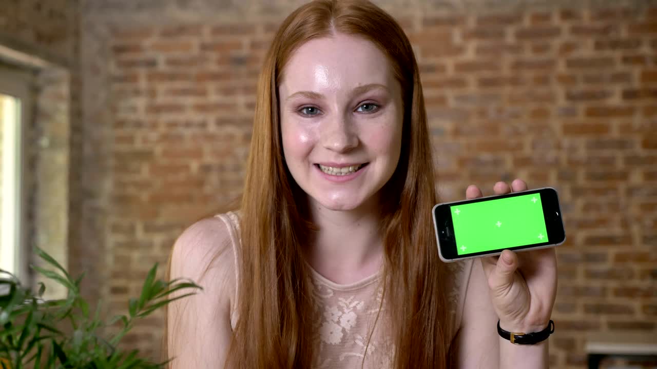 Young fresh ginger girl is showing green screen on her smartphone, watching at camera, communication concept, brick background