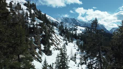 Panorama-drone-fly-through-trees-at-scenic-Schachen-near-Bavaria-Elmau-castle-away-from-the-snowy-glacier-mountain-tops-in-the-alps-on-a-cloudy-and-sunny-day-along-rocks,-forest-and-hills-in-nature