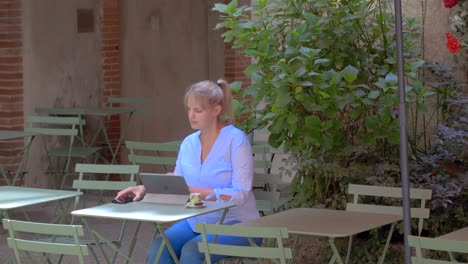 A-blonde-Caucasian-woman-with-her-hair-tied-back,-sitting-on-a-café-terrace-using-her-tablet,-smartphone-and-having-her-coffee