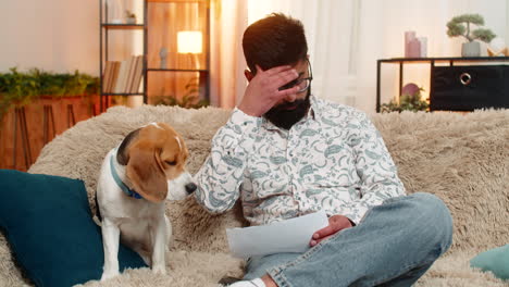 indian man sadly checks utility bills while beagle dog shows emotional support on sofa at home