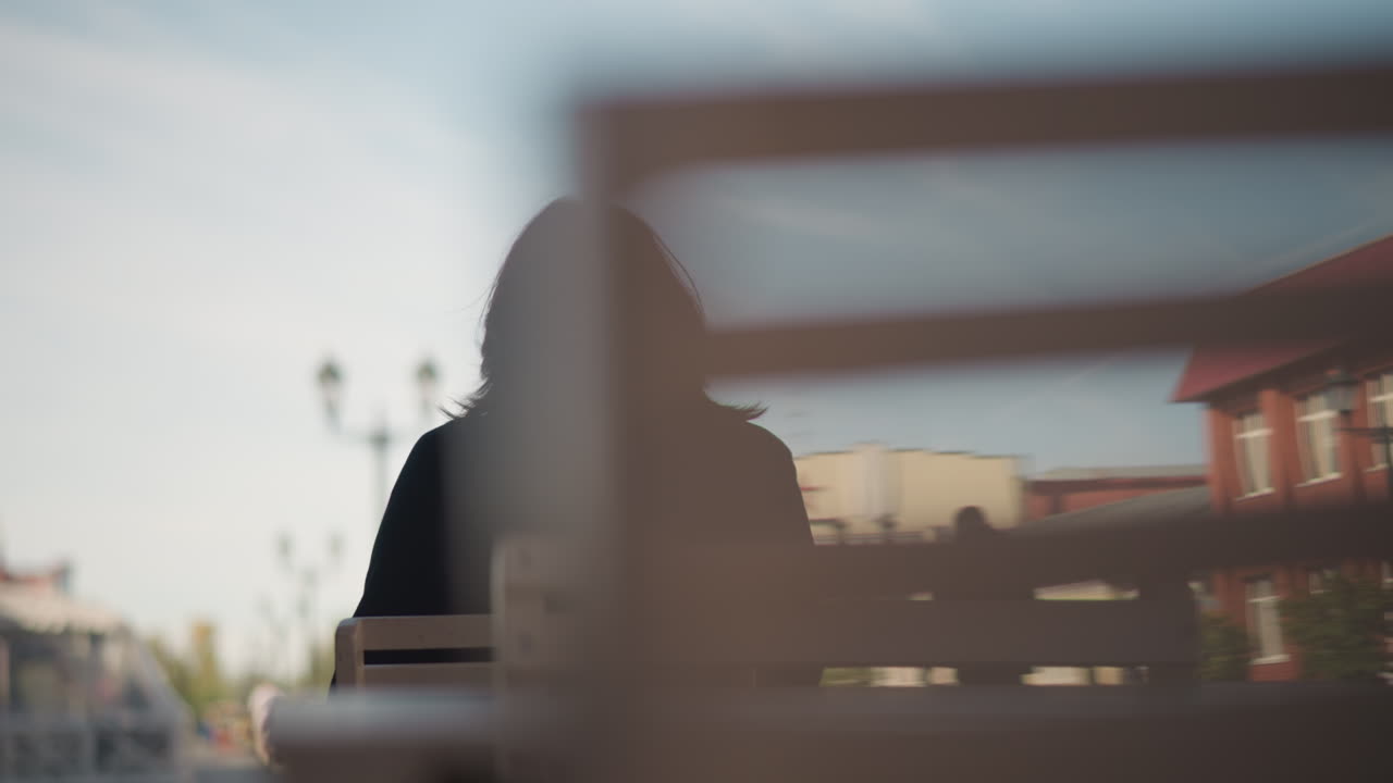 은 하늘 아래 먼 건물과 거리 조명에 직면한 노트북에서 일하는 열린 도시 환경에서 야외에서 나무 의자에 앉아있는 여인