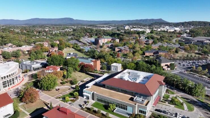 Volo di drone sopra la James Madison University ad Harrisonburg, con cortile interno ben curato ed edifici storici. Alberi colorati e cielo blu con montagne in lontananza. Ripresa aerea. Campo largo