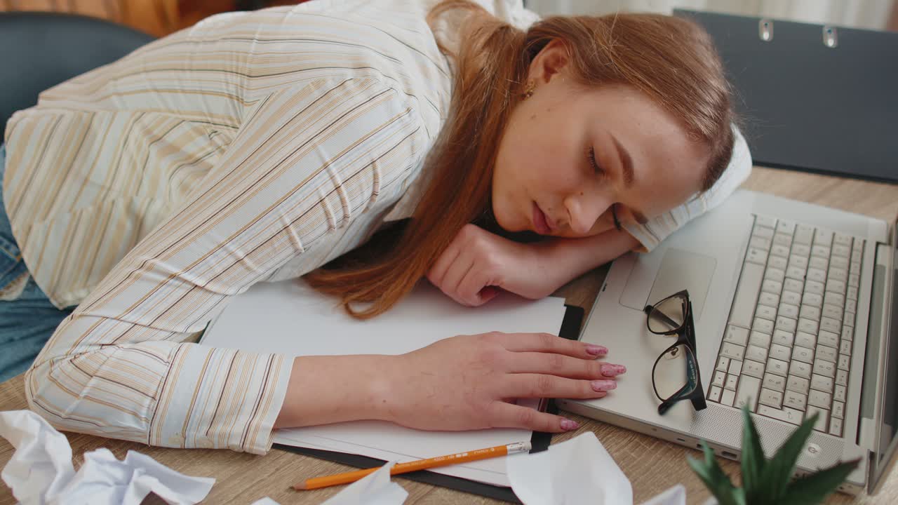 Tired woman at home office falling asleep on table with laptop computer crumpled sheets of paper