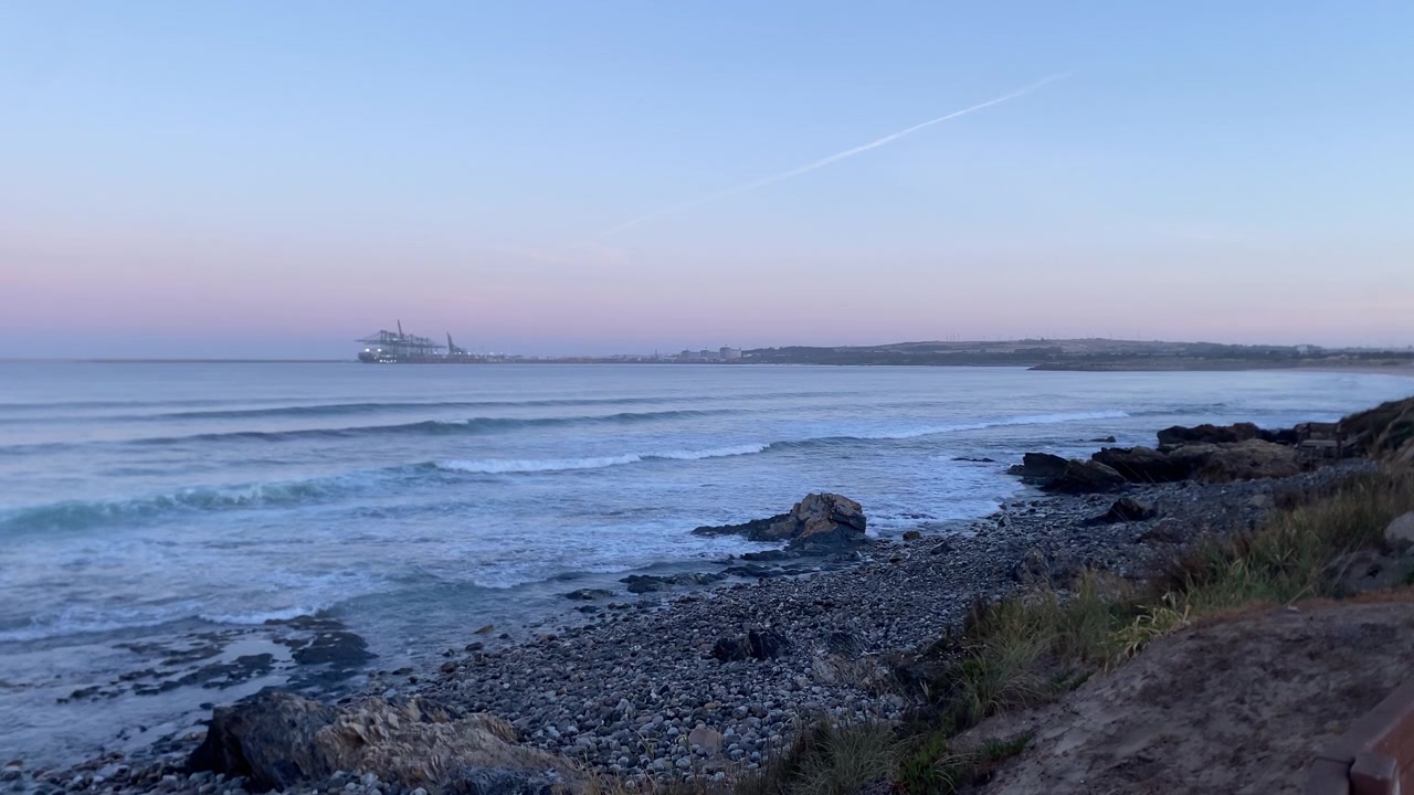 Alentejo Portugals kystlinje med blide havbølger, der flyder ind på en stenstrand under en fredfyldt skumring, med udsigt til en fjern havn.