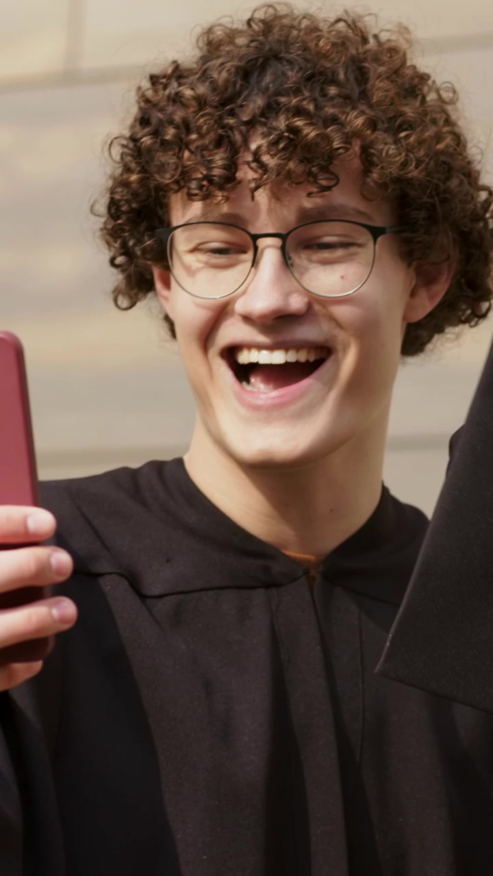 Video verticale: Tenendo un telefono rosso, un laureato con felpa con cappuccio reagisce alla laurea davanti a un muro, tenendo un tocco nero