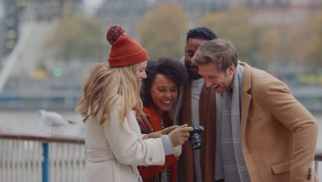 group of friends outdoors wearing coats and scarves looking at photos on camera on autumn or winter trip to london - shot in slow motion