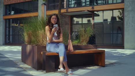 Asian-brunette-sitting-with-phone-on-city-bench.-Girl-using-mobile-gadget.