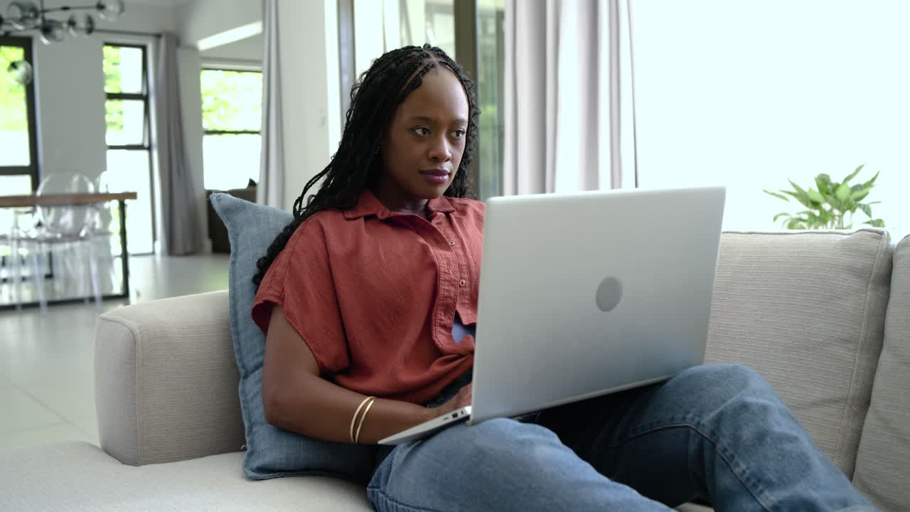 African woman typing on silver laptop on lap at home, pausing while reading screen for work