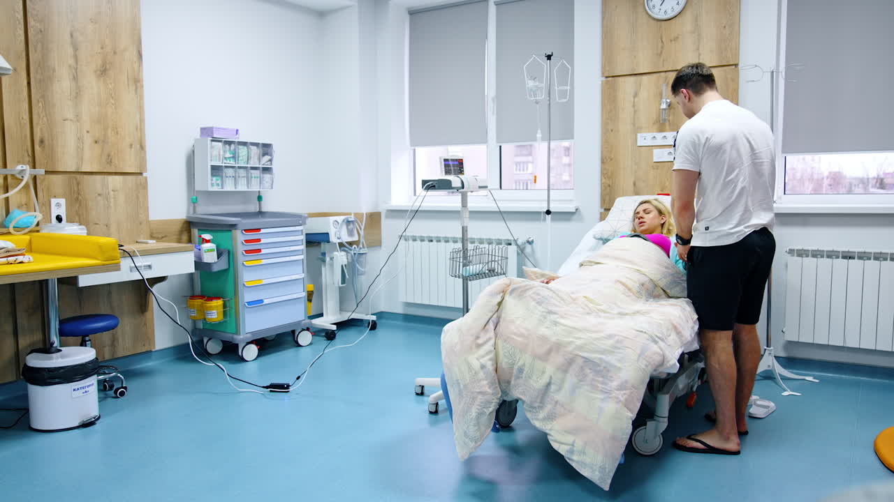 Woman feeling painful contractions lies in the hospital bed. Man standing beside comforts her stroking her head.