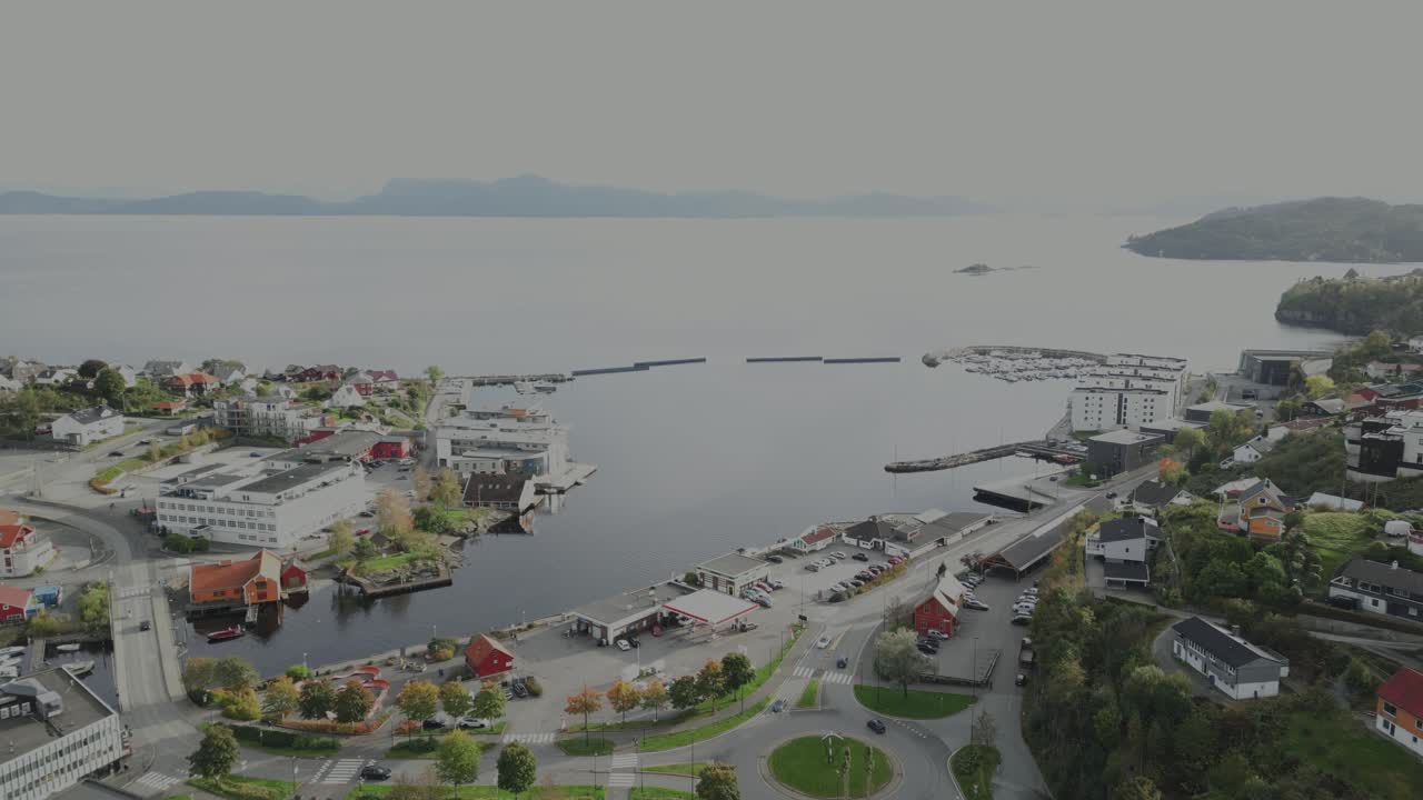 This video shows a town by a fjord, with a harbor, colorful houses, and mountains in the distance. The view is from a high angle