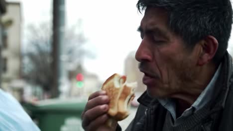 a man sits near a dumpster, savoring a piece of street food. he appears contemplative as he enjoys the meal in a bustling city environment during cool weather.