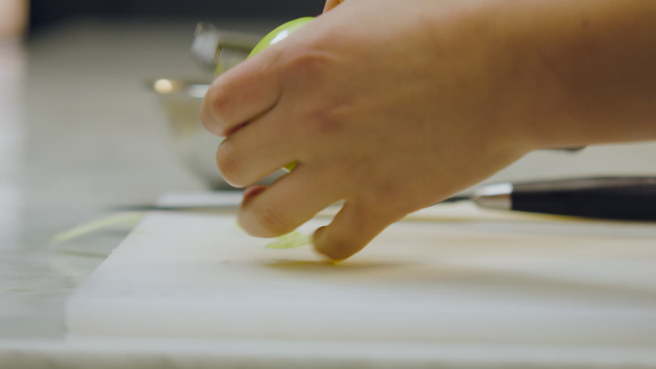 Close-up of a chef peeling a green apple with a peeler, while in the out-of-focus background a knife and an aluminum mold can be seen