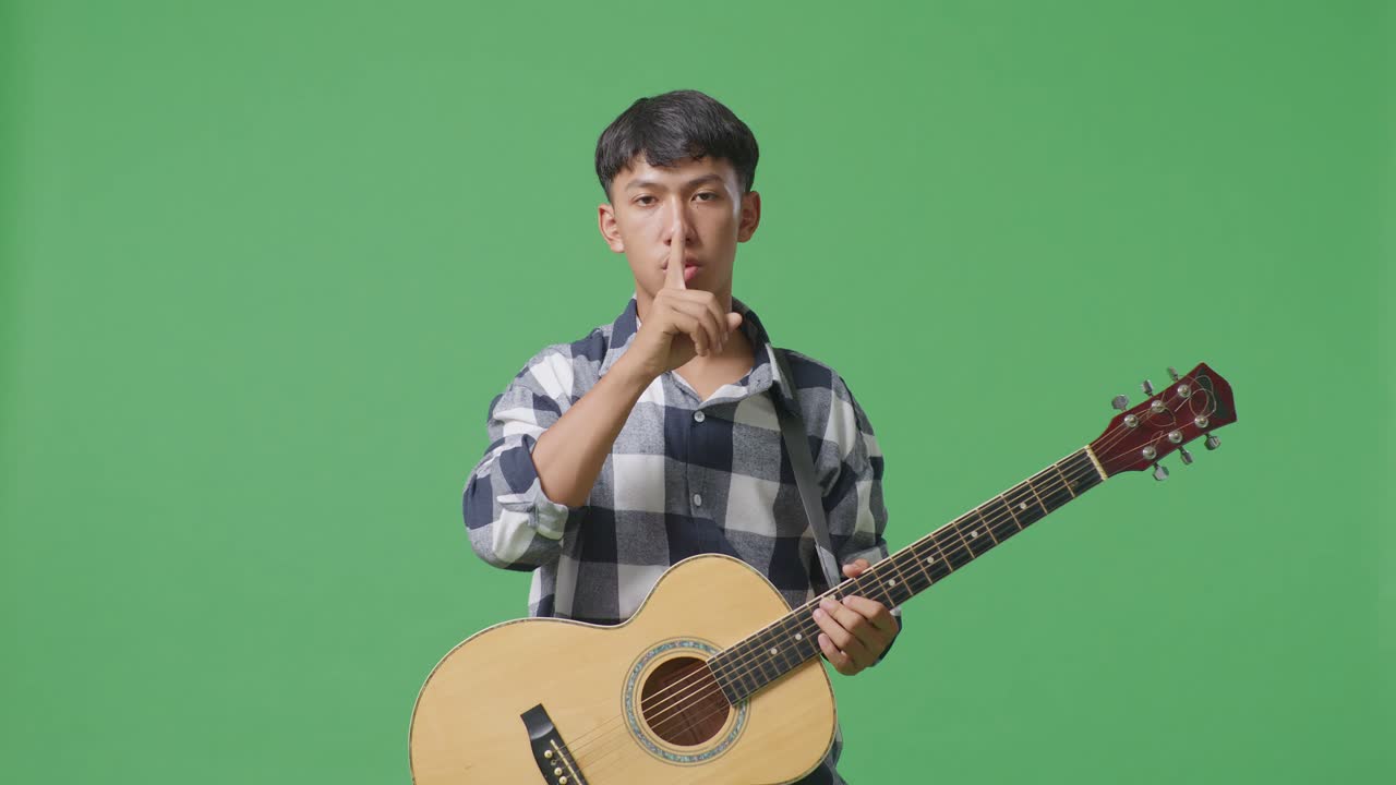 초록색 스크린 배경 에 서 있는 동안 기타 를 들고 있는 아시아 청소년 이 침 의 몸짓 을 보여 준다