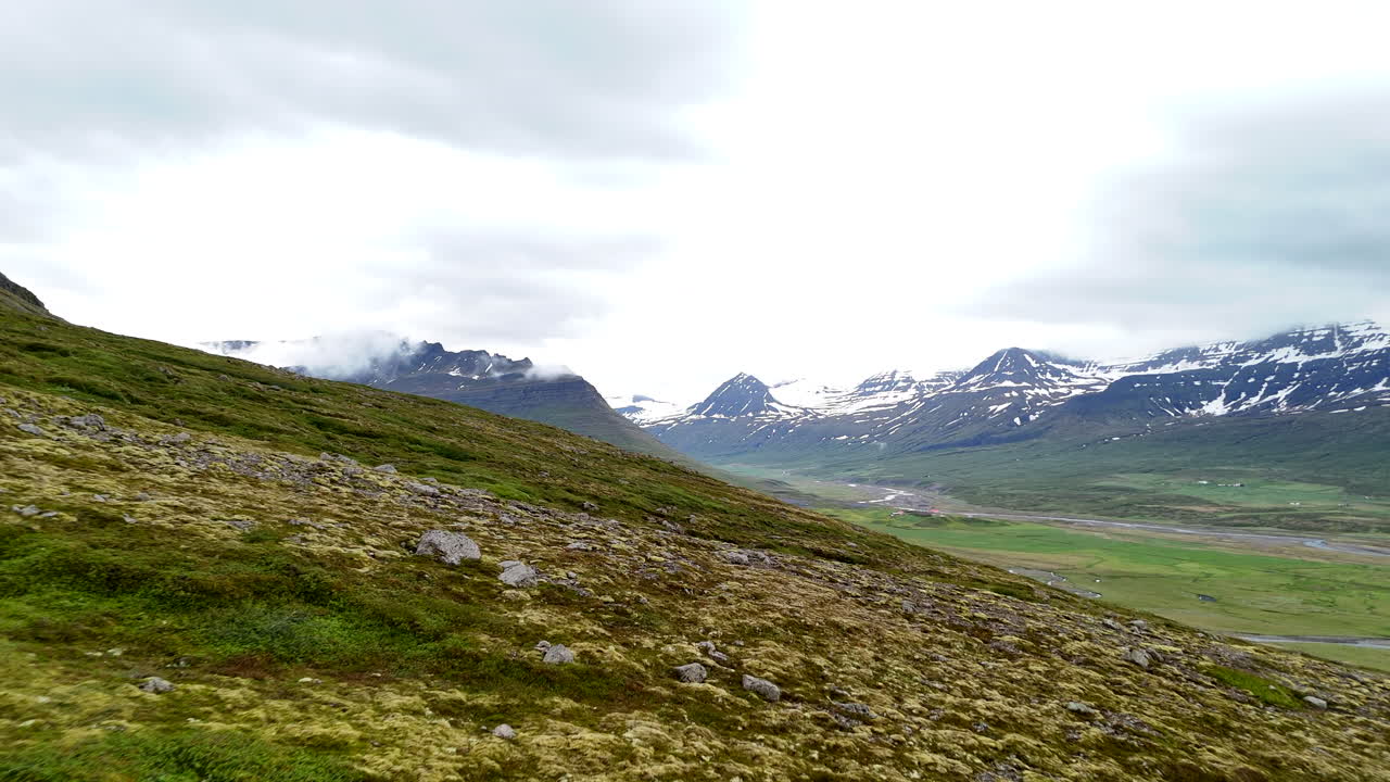 Wide aerial view of the mountains near Fáskrúðsfjörður, with snow-lined peaks, scattered waterfalls, winding wetlands, and an open valley crossed by a curving road under cloudy skies
