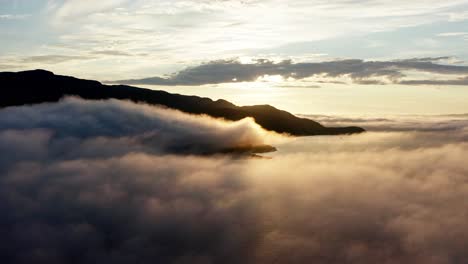 drone flying above the clouds over the sea with rocky mountain in silhouette on a golden hour sunrise - wide aerial shot