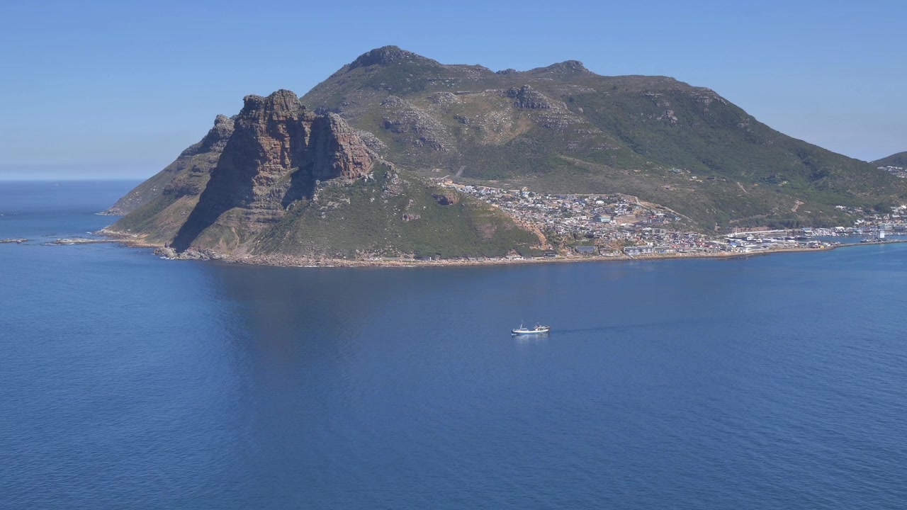 Dall'alto, un peschereccio a strascico esce dal porto di Hout Bay nell'oceano sconfinato, con un'imponente montagna che si erge sullo sfondo.
