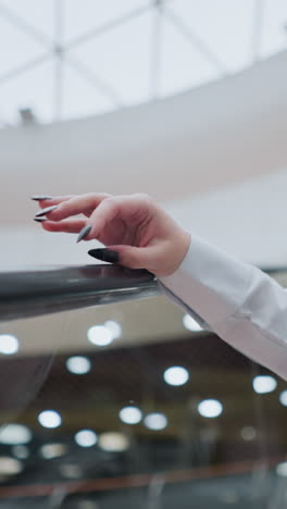woman with black polished nails gently taps escalator handrail while descending, she wears a white blouse and jeans, soft-focus background