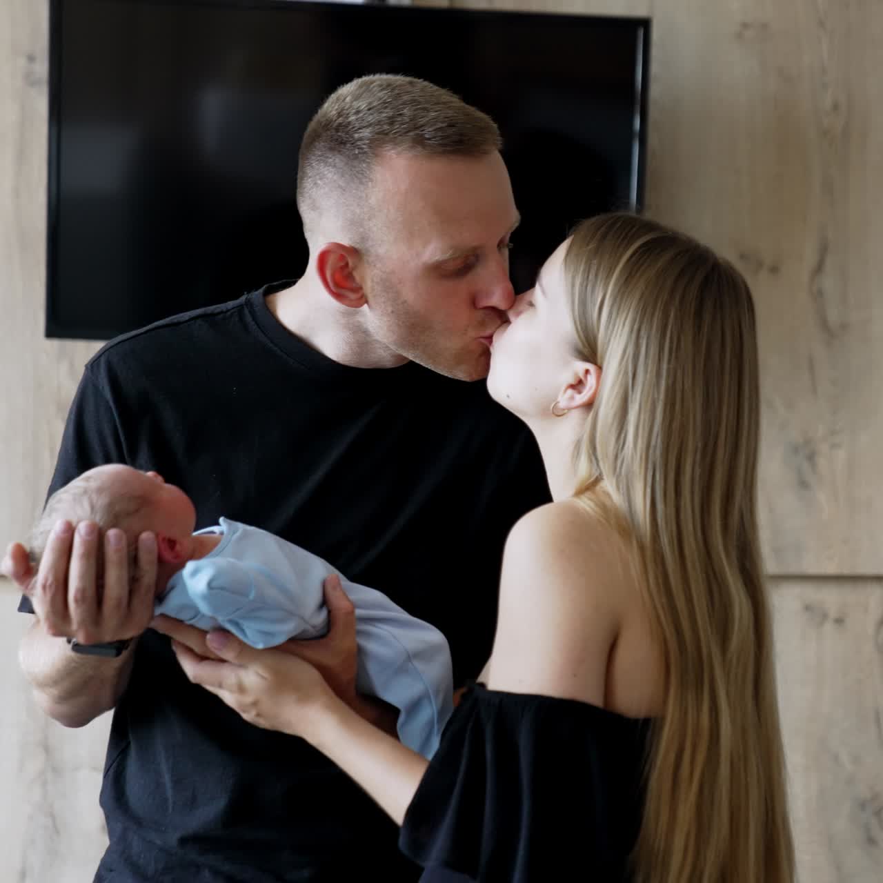 Man is holding his newborn son a kissing his wife standing beside. Happy new-made parents with their child