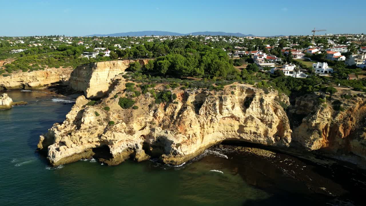 Drone flying above coastal cliffs near Faro, Portugal showing golden rock formations, shallow waters, trees, houses with red roofs, winding shoreline, and mountain range visible in distance