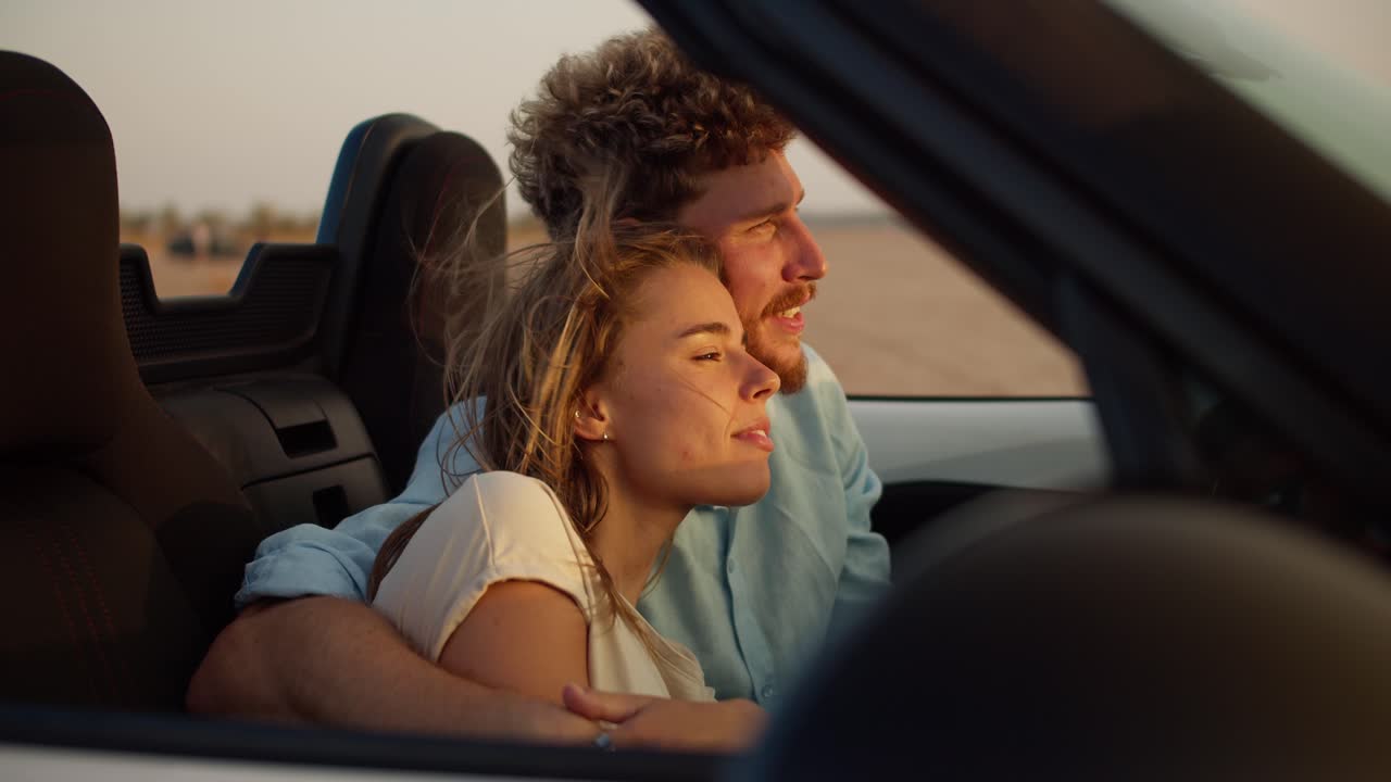 A Bearded Guy With Curly Hair Hugs His Blonde Girlfriend And Caresses Her.  A Guy And A Girl Are Sitting In A Convertible Car In Windy Weather On The  Field Free Stock