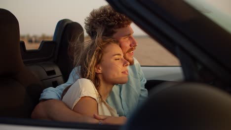 A-bearded-guy-with-curly-hair-hugs-his-blonde-girlfriend-and-caresses-her.-A-guy-and-a-girl-are-sitting-in-a-convertible-car-in-windy-weather-on-the-field