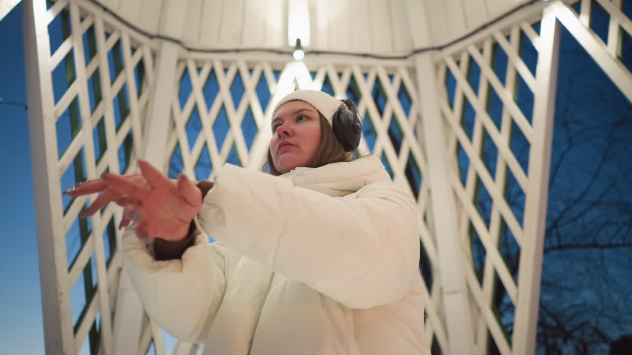 Young lady wearing white puffer coat and knit beanie moves hands while listening through headphones inside illuminated latticed pavilion at dusk with snow outside and overhead lights adding warm glow